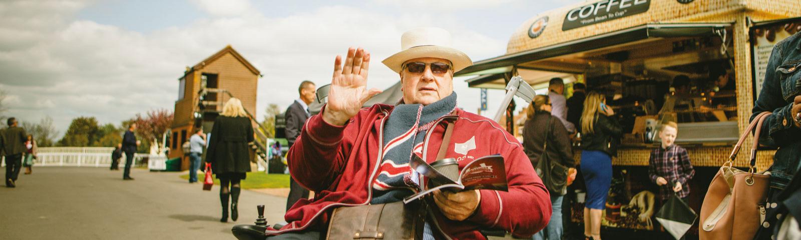 A racegoer waves for the camera as he makes his way in his wheelchair past a mobile coffee van.